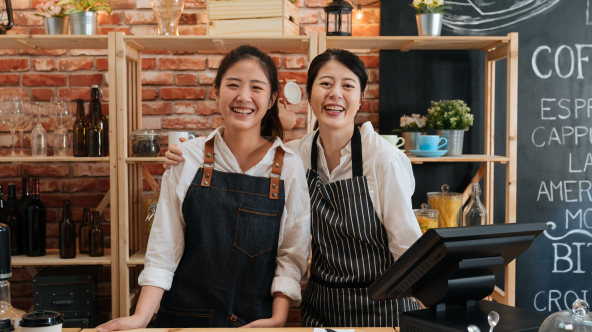   A female with her arm around another female both wearing aprons in a shop