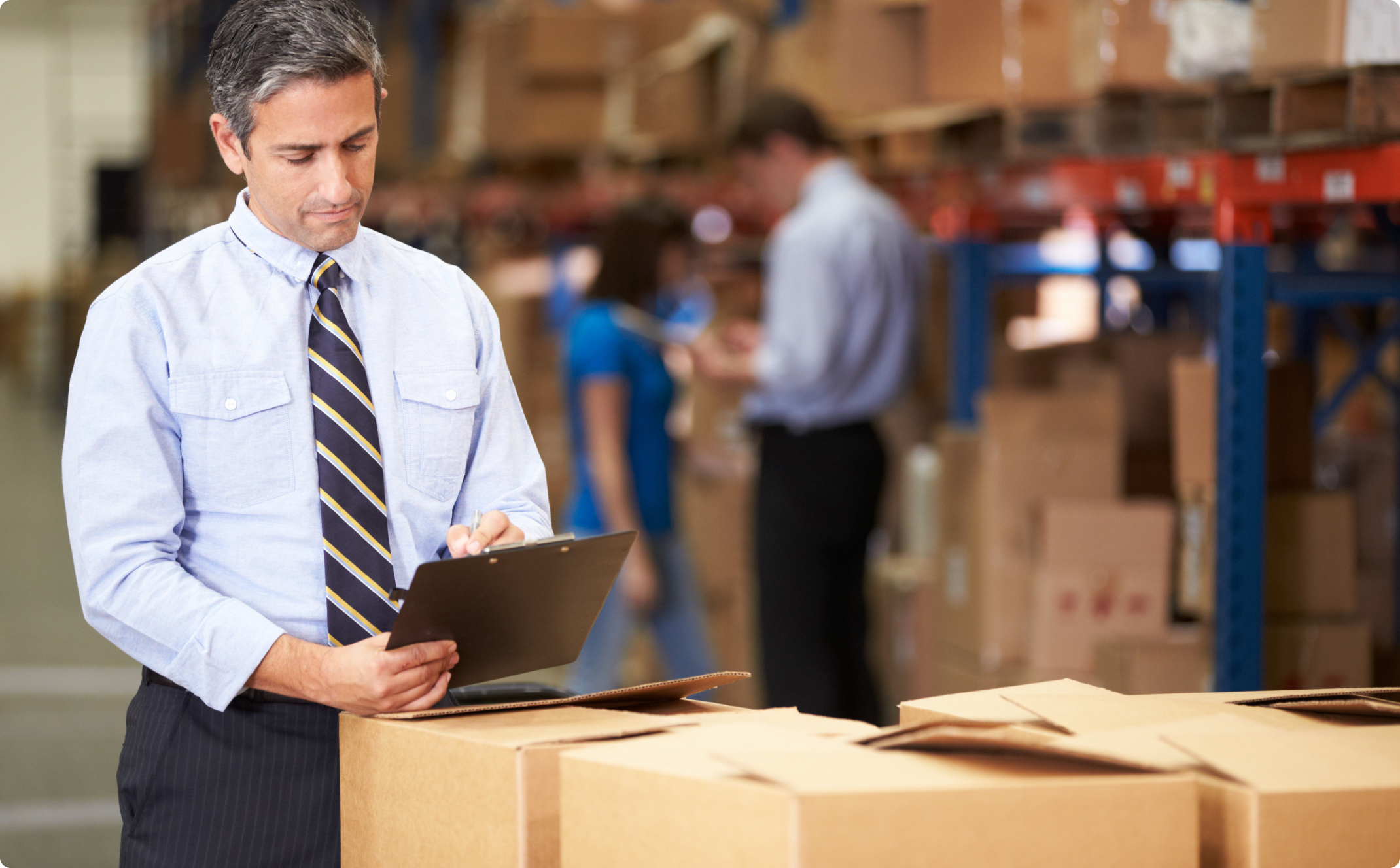 Male writing on clipboard in warehouse full of boxes